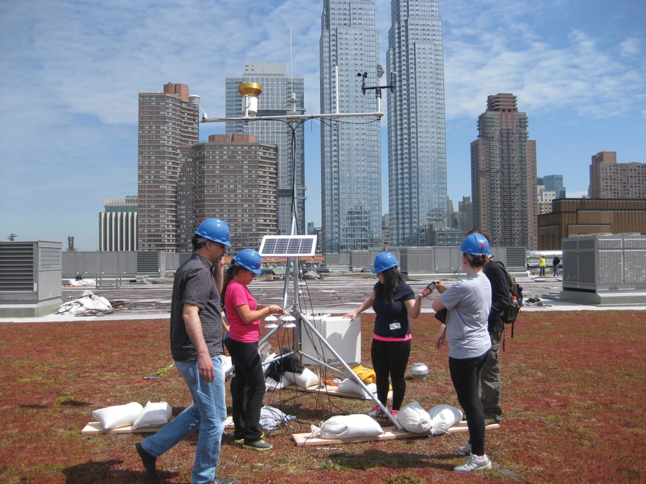 A group of people with white bags of equipment and electrical wiring standing on top of a green roof with a metal structure
