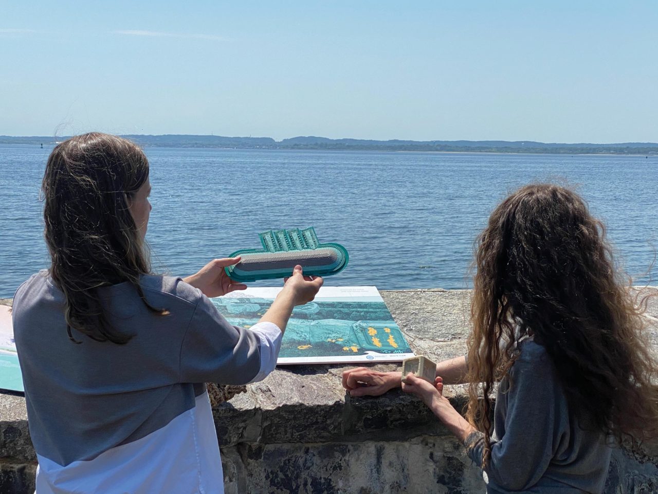 Two women looking out over the water and holding a small plastic or metal model and looking at a map of the sea floor