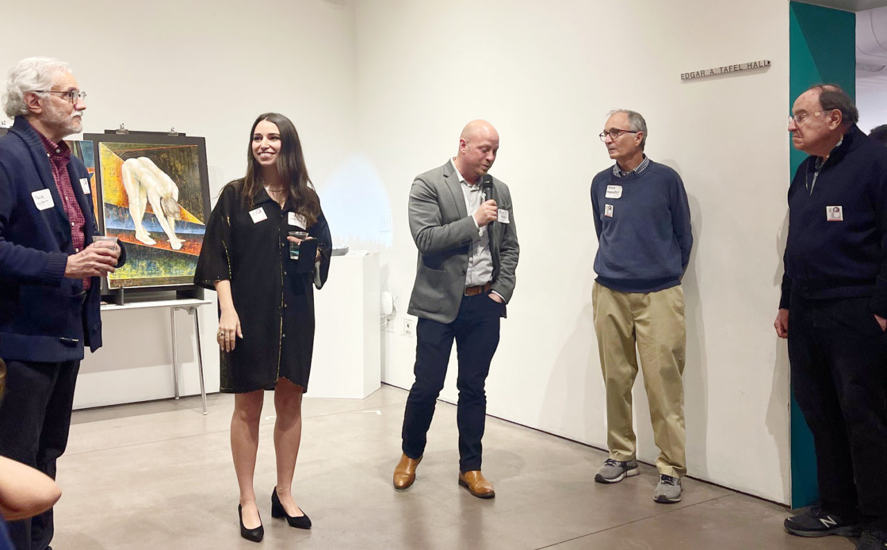 Five individuals, one holding a microphone, lined up inside a room with a painting behind them.