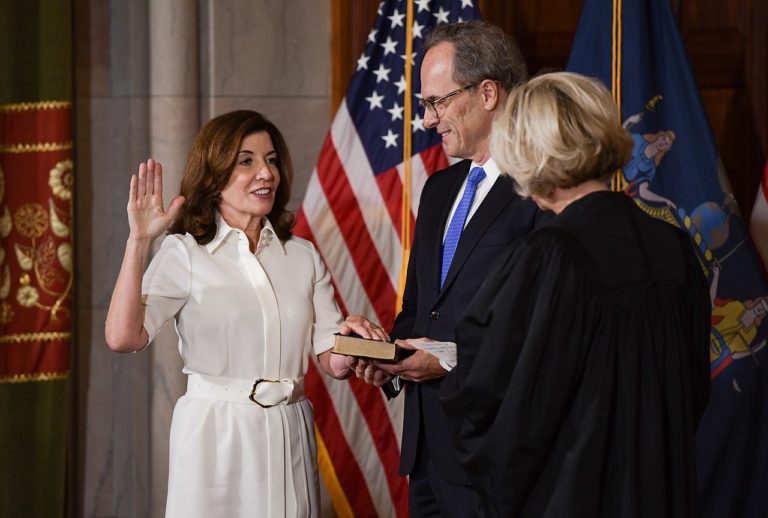 Woman in white dress raises right hand while putting left hand on top of a book held by a man.