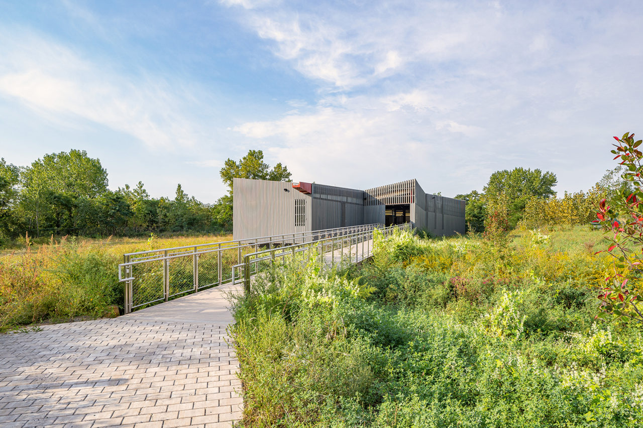 A grey brick path leads to a grey structure with vertical striations set against marsh grasses