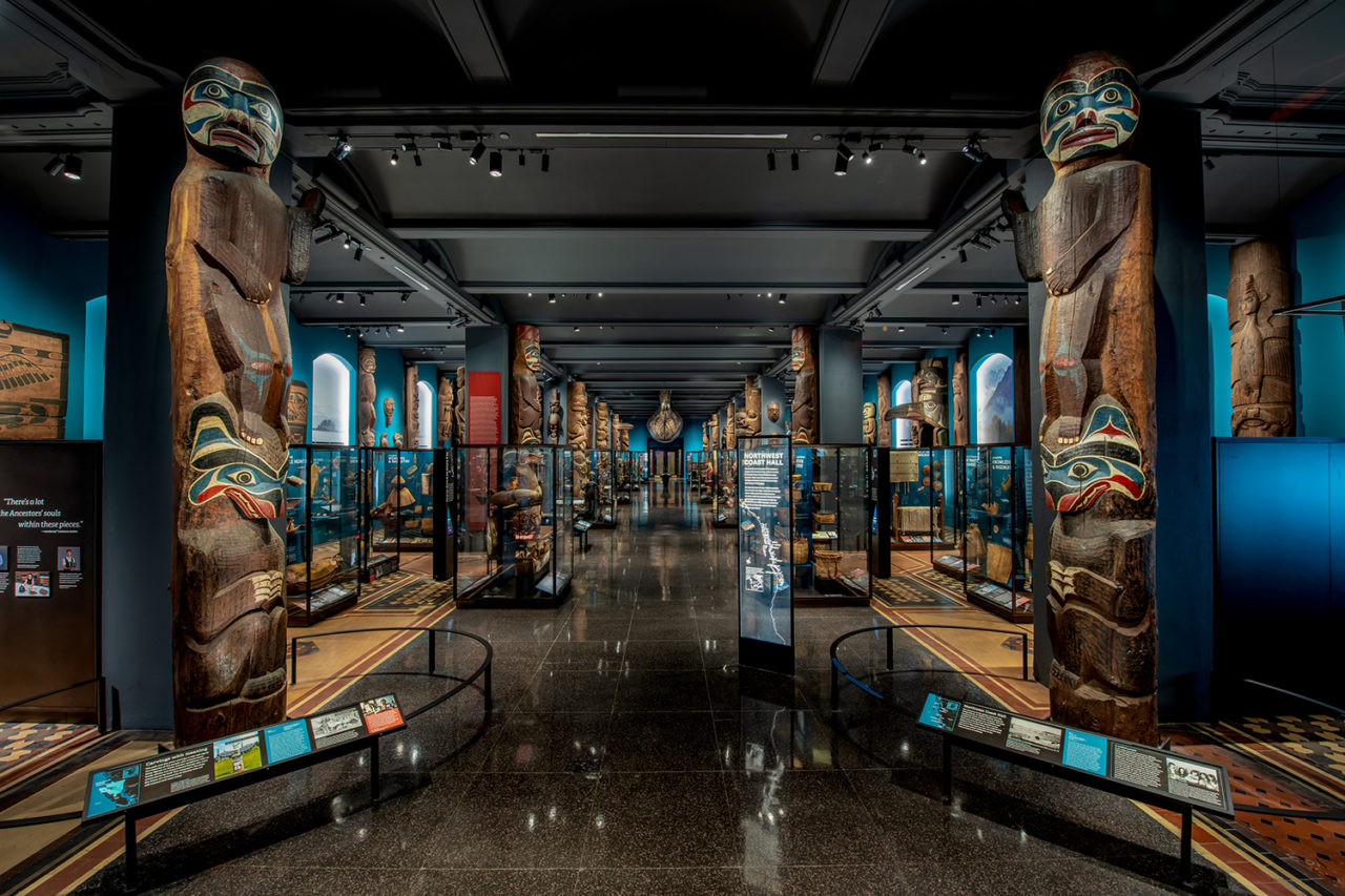 View down a spacious exhibition hall featuring large, wood-carved artifacts and teal walls