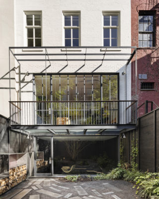 The exterior of a townhouse with a second floor balcony that is surrounded by a steel structure. On the ground level there are green plantings to the right and stacked, chopped wood to the left