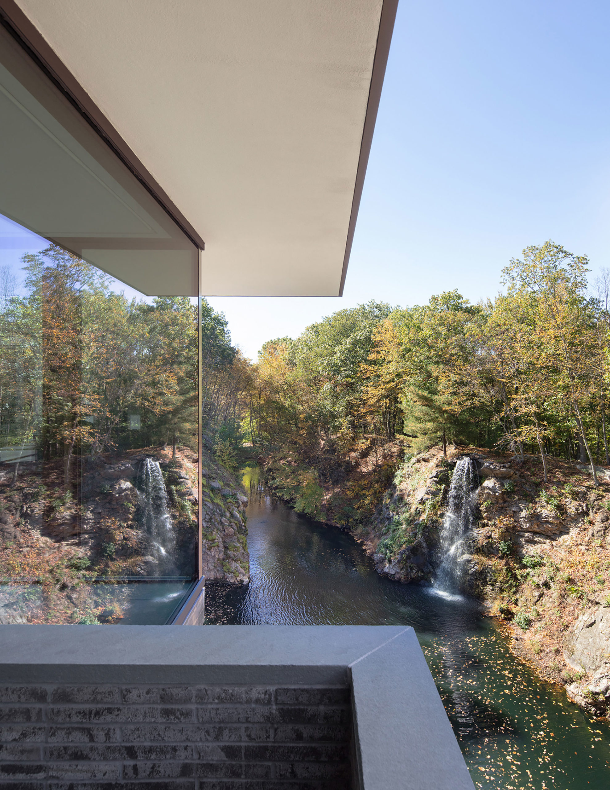 A closeup on the exterior of a house with a glass wall reflecting a waterfall and a leafy hillside