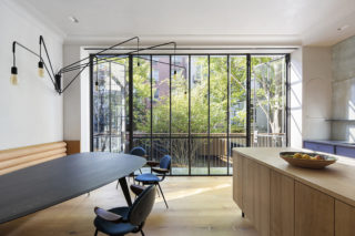 A home interior with a dining table to the left and a wooden counter to the right with a bowl of citrus on top. A glass door looks out over a balcony with more apartments and trees outside