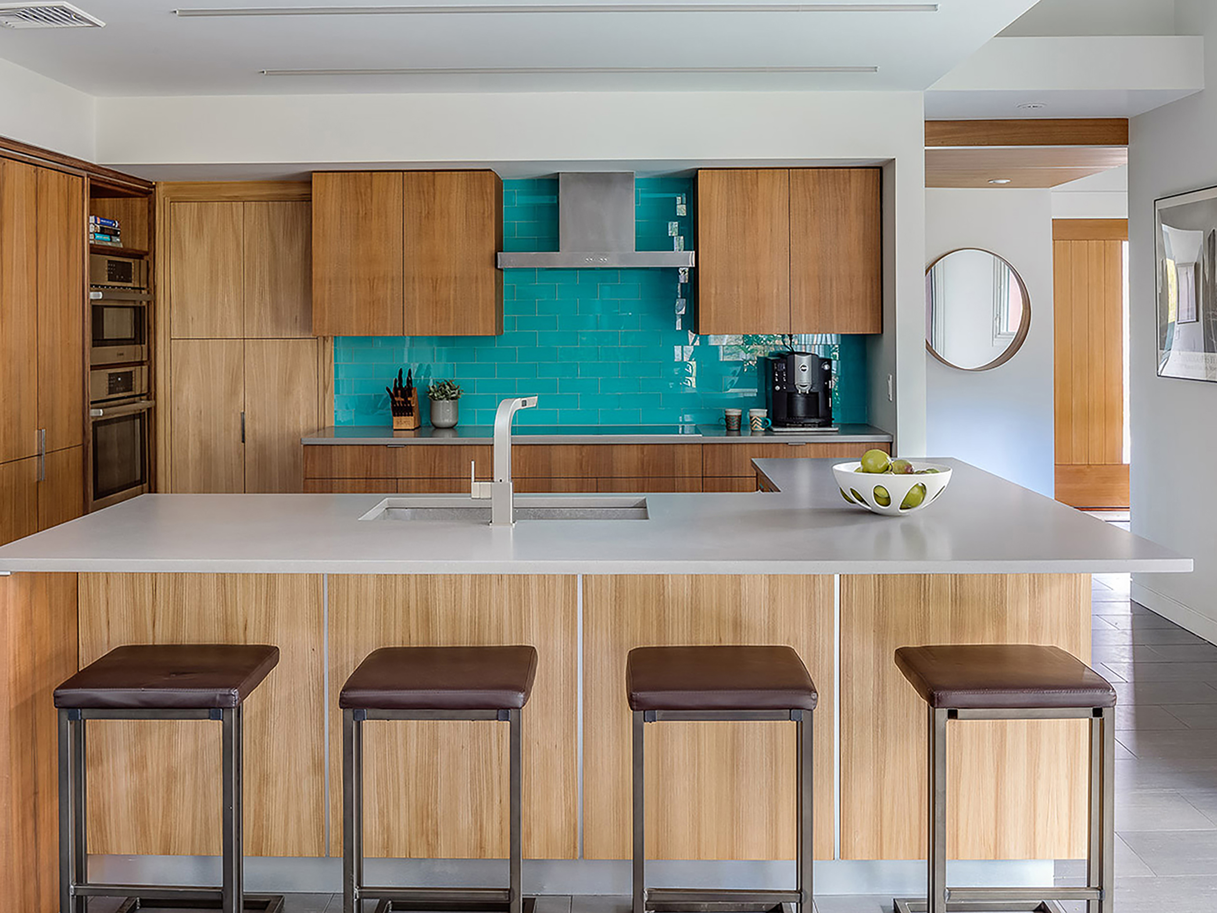 A kitchen counter with a sink and four bar stools in front of it, with wooden cabinets and a green backsplash in the background
