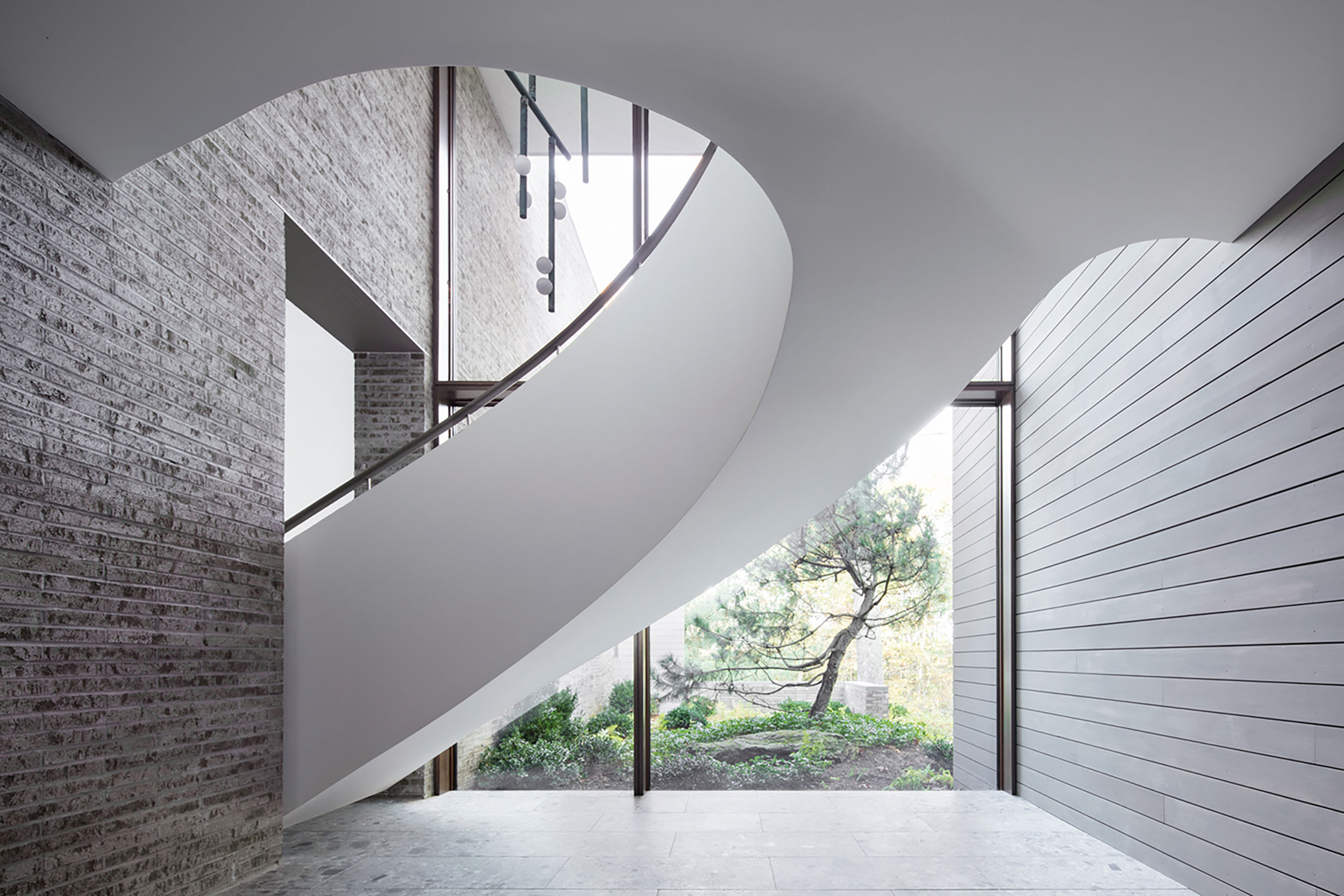 The smooth, white underside of a staircase suspended over a stone floor, with a glass wall in the background looking out to nature