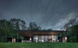 The exterior of a house at dusk or dawn, with floor-to-ceiling windows on the front and a green lawn