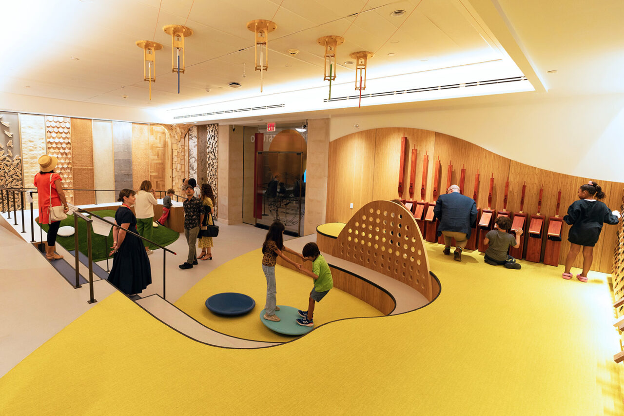 Children and their caregivers are scattered in the 81st Street Studio, a wide play room in The Met with a yellow painted floor and tactile wooden surfaces seen in the back.