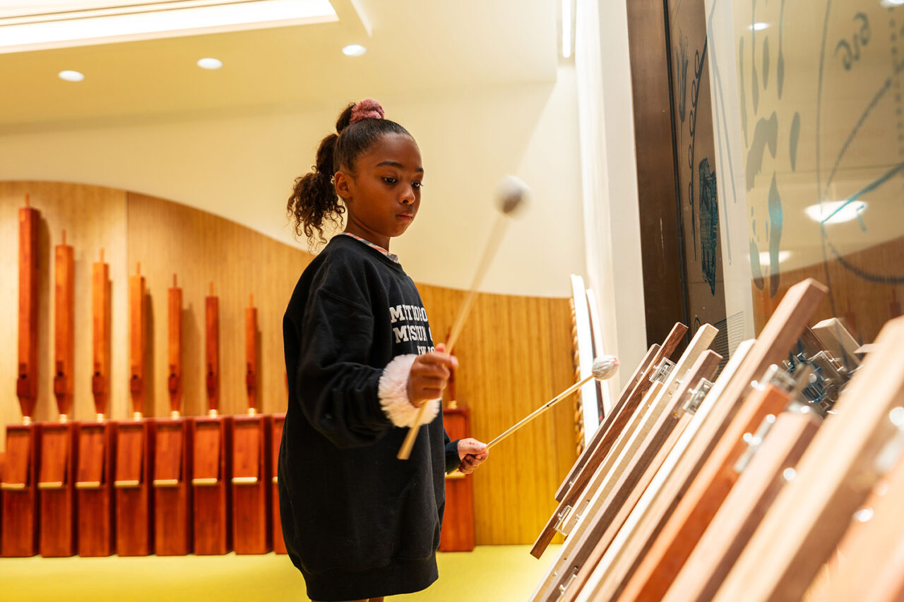 A young Black girl hits a wooden xylophone inside the 81st Street Studio.