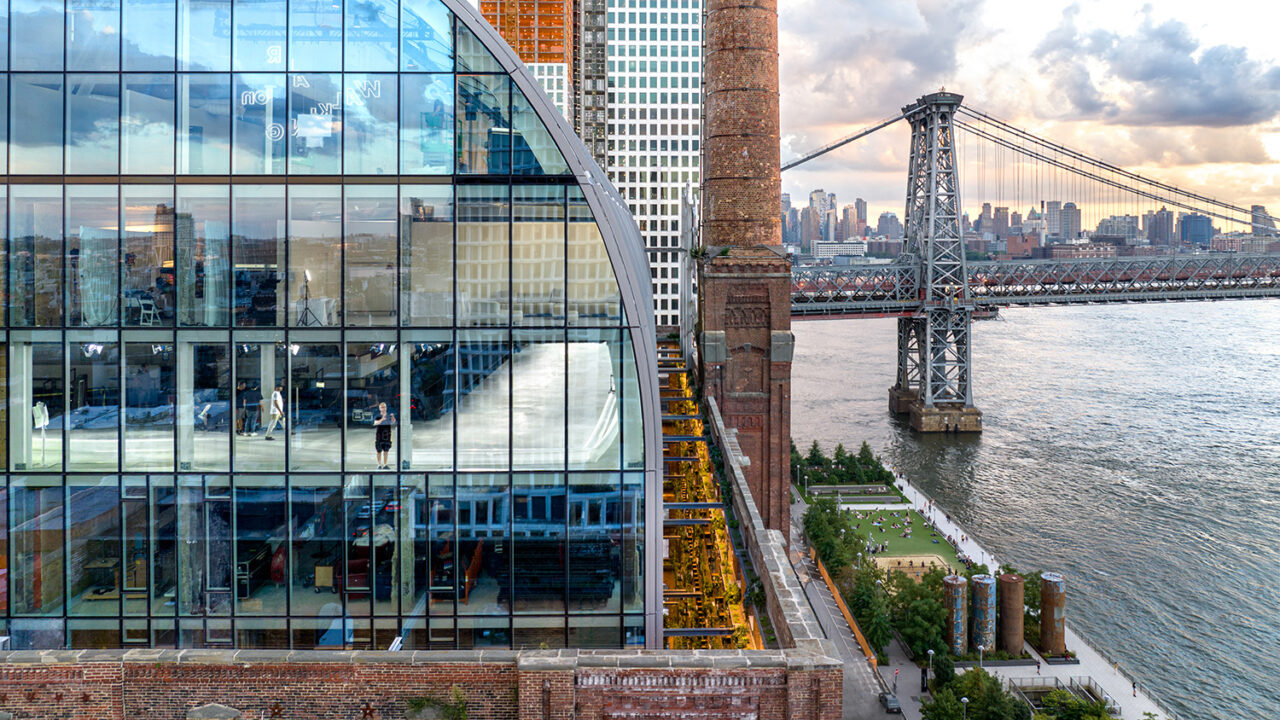 A digital rendering of people in the glass rooftop area of the office building. The Williamsburg Bridge is in the background.