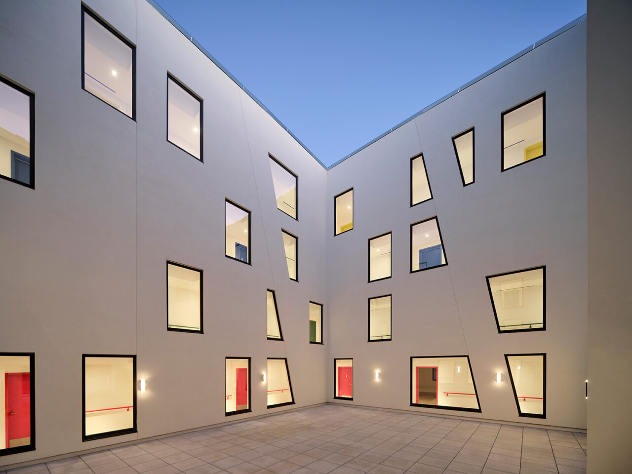 The view of an apartment complex from the central courtyard. The walls are white with windows in a variety of sizes that show corridors and colorful doors.