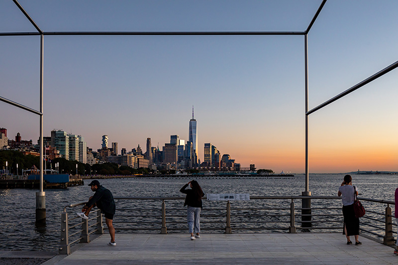 Gansevoort Peninsula: people standing on a structure above the water with a city scape in the distance.