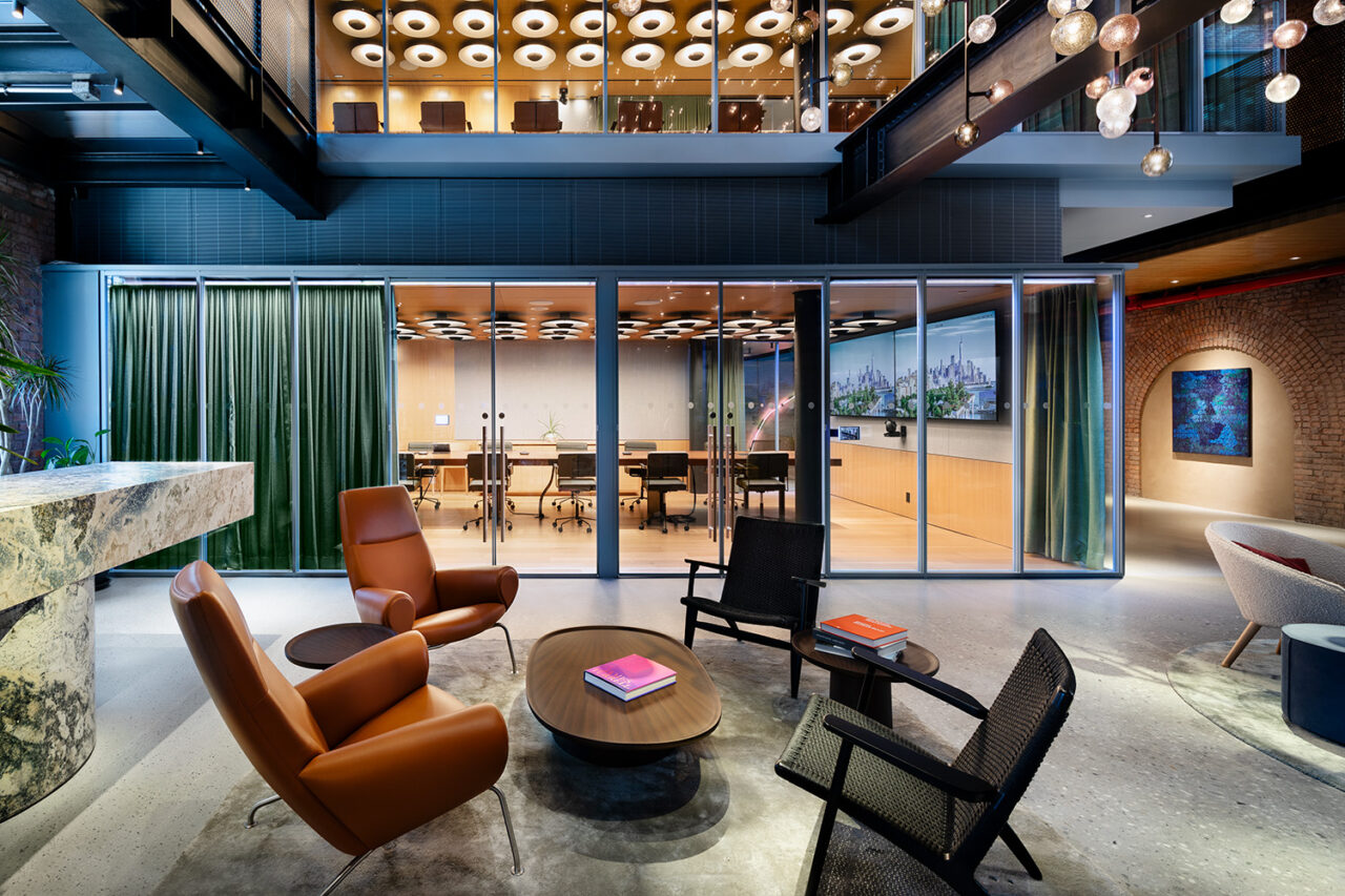 The central gathering area in an office. Four chairs sit around a low-to-the-ground circular desk. A warmly lit meeting room is in the background.