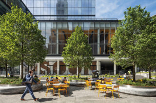 A street view photograph of some yellow tables and chairs in the courtyard by a glass building.