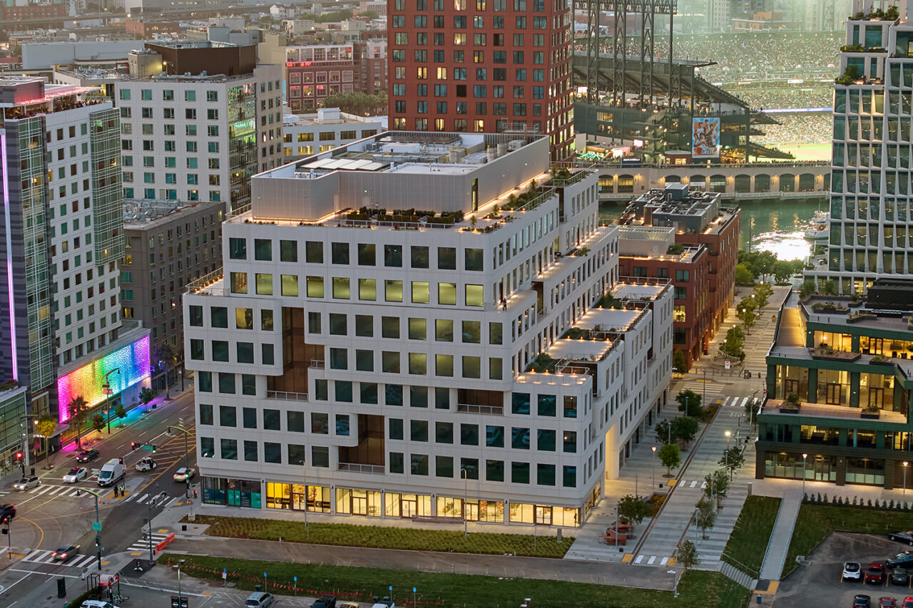 Aerial view of a white building with dark windows all over. The structure is made up of various layers and levels. A sports stadium and various city buildings in the background.