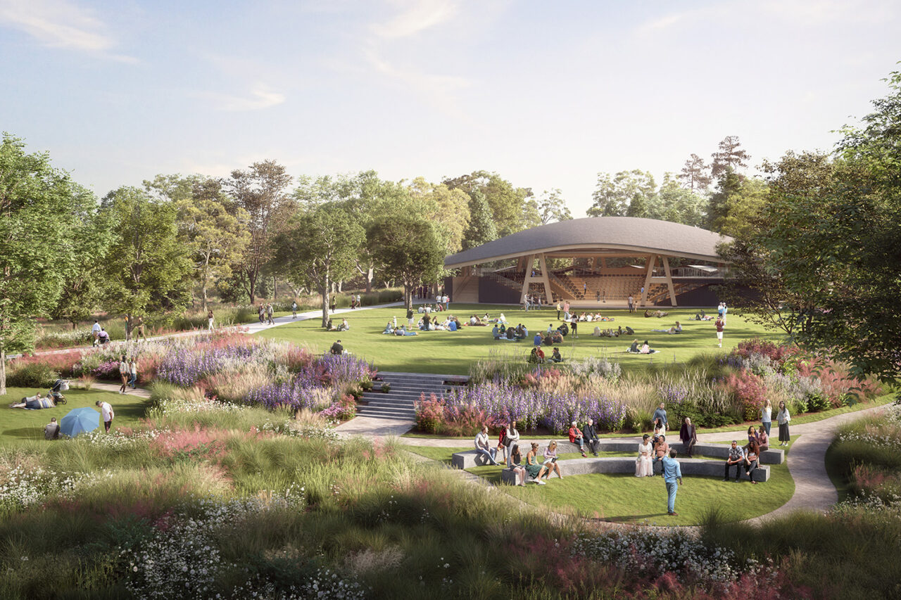 A landscape view of people sprawling around the green, with paths leading up to the theatre in the distance. Trees and greenery surround the scene.