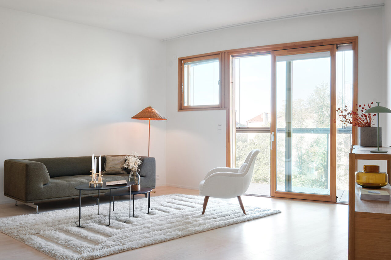 An interior view of a small living room, with a white chair, a gray couch, and orange lamp. Light streams in through the large windows.
