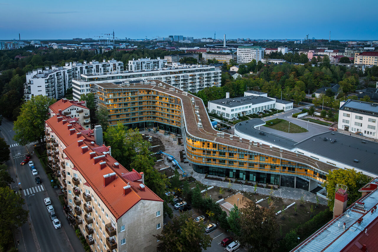 An aerial view of a large glass building with a smooth curved shape. Classic apartment buildings surround the dimly lit structure.