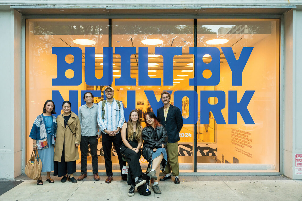 The Once Future graphic design team poses in front of a warmly lit Center for Architecture. Blue text "Built by New York" is displayed on the glass panel behind them.