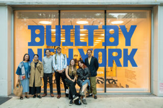 The Once Future graphic design team poses in front of a warmly lit Center for Architecture. Blue text "Built by New York" is displayed on the glass panel behind them.
