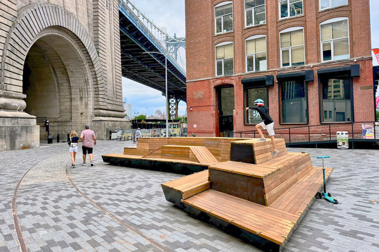 View of a sculptural wooden bench with the Brooklyn Bridge in the background.