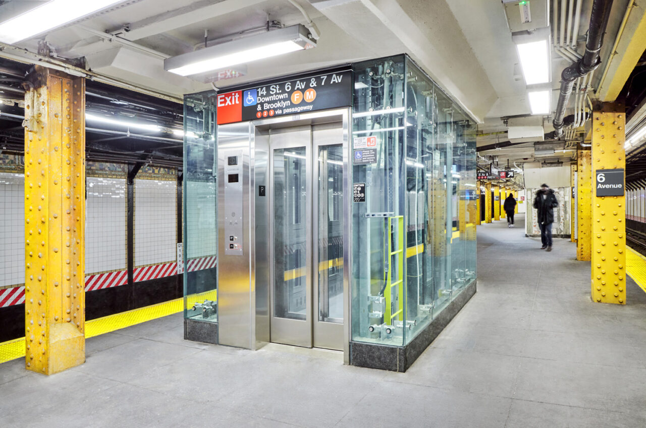 An elevator made of steel and glass at the 14th street 6 Av & 7 Av subway station.