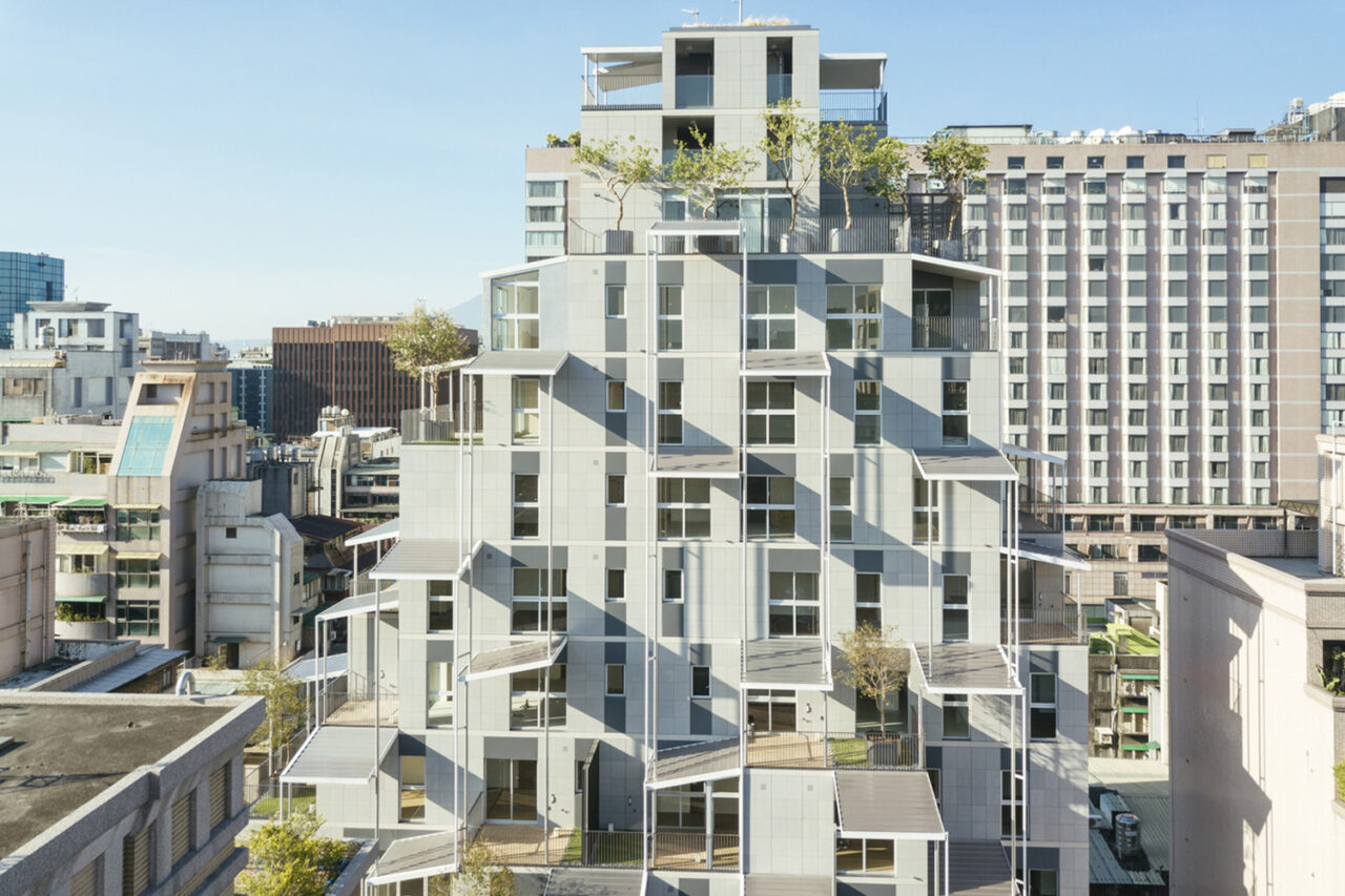 A light blue-gray colored building, with geometrically stacked units and trees on balconies.