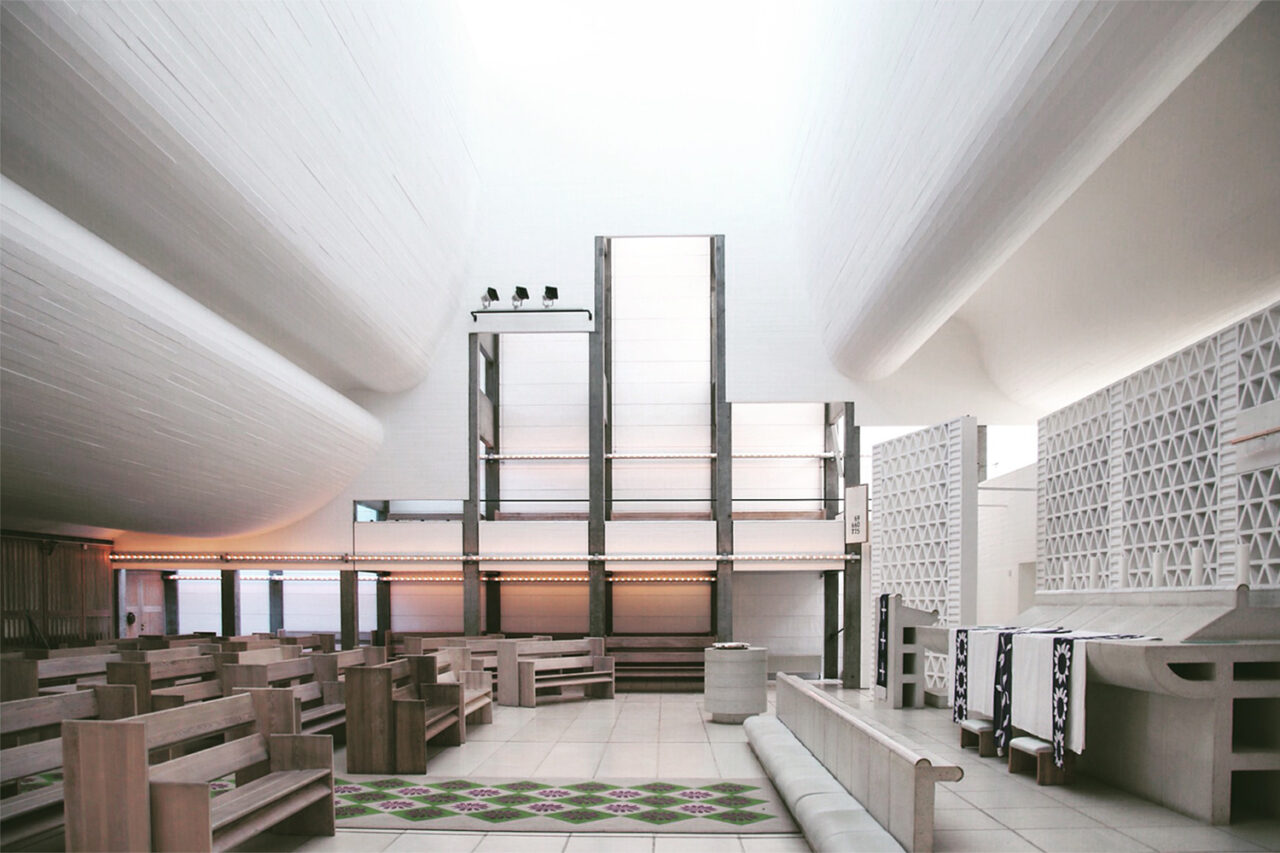 Interior scene of a white curved, sculptural ceiling. Rows of wooden benches on the left, an altar scene on the right.