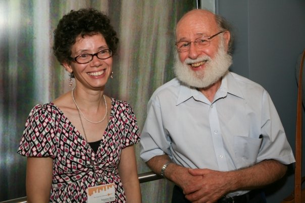 (Left) A white woman with short curly hair and glasses, smiling alongside (right) an older white bald man with a white beard.