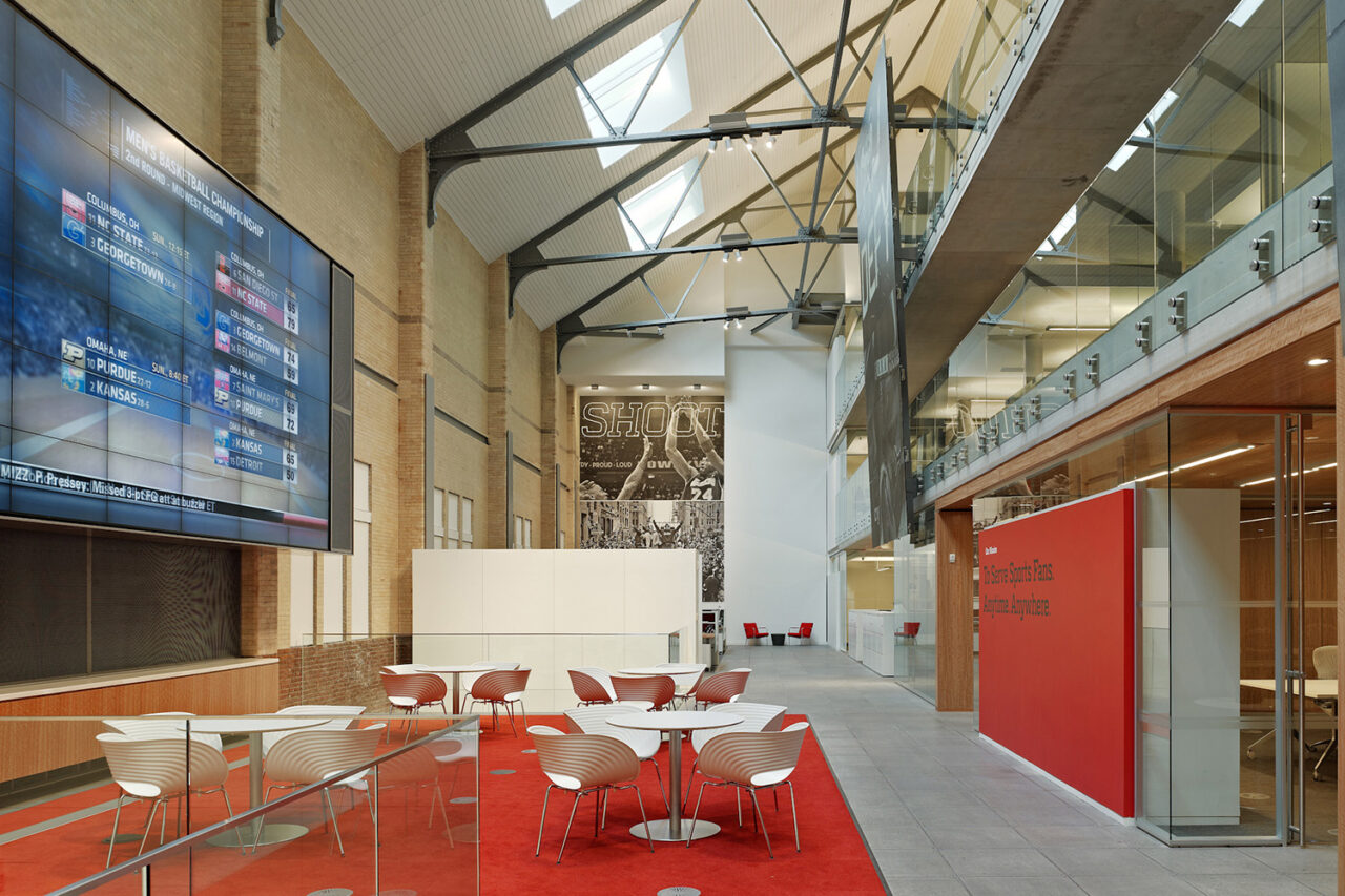 Interior shot of a room with tables and red carpet. A large screen on the left. High ceilings and skylights in view.