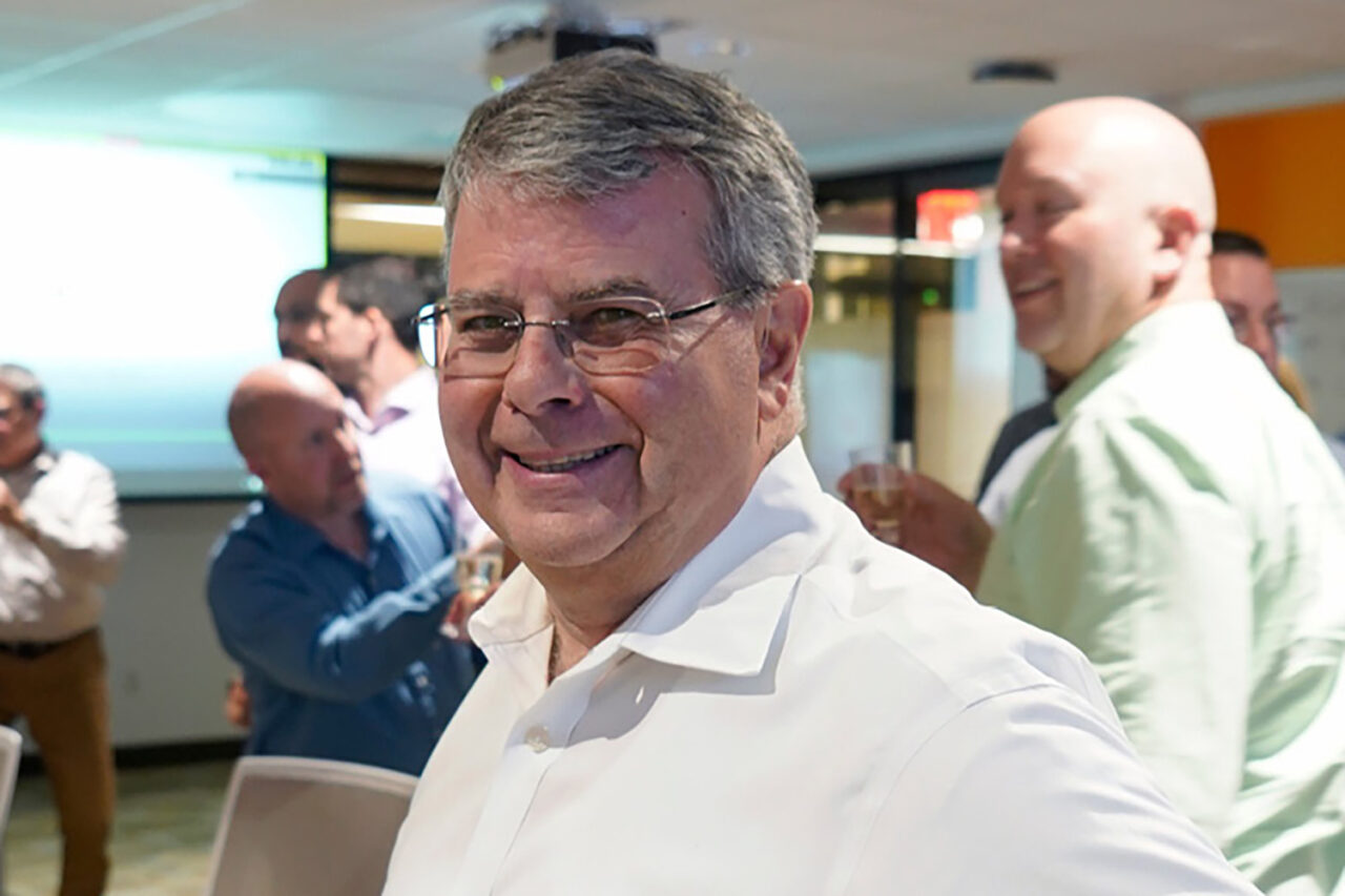 A white man with silver hair wearing glasses smiles at the camera. He wears a white collared shirt.
