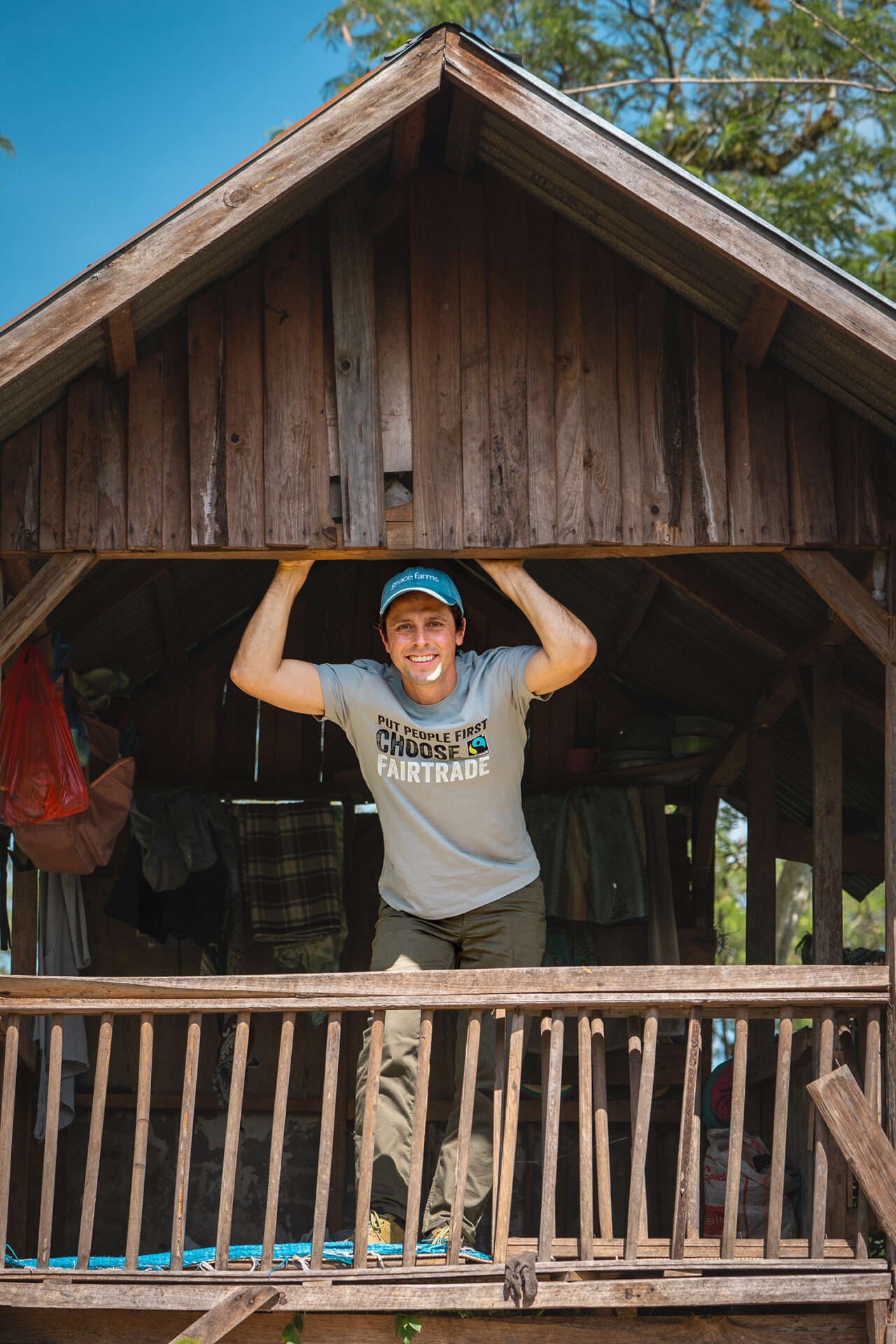 White man in gray shirt and blue cap smiles from the balcony of a brown cabin.