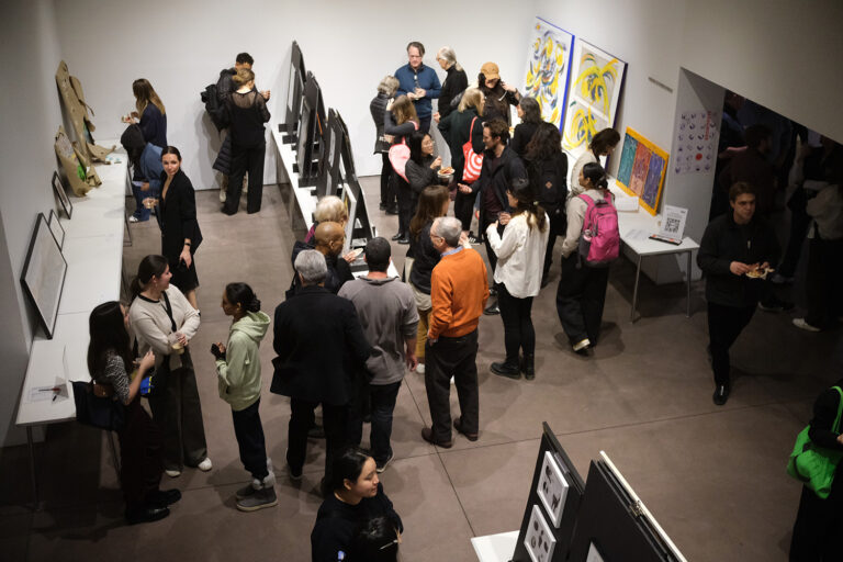 A birds eye view of the Art by Architects showcase installed in a gallery setting with people walking around looking at art