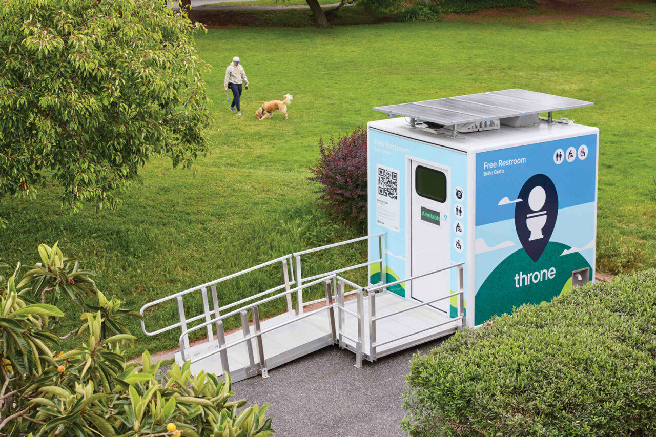 A modular restroom in font of a grassy area where a person and a dog are playing
