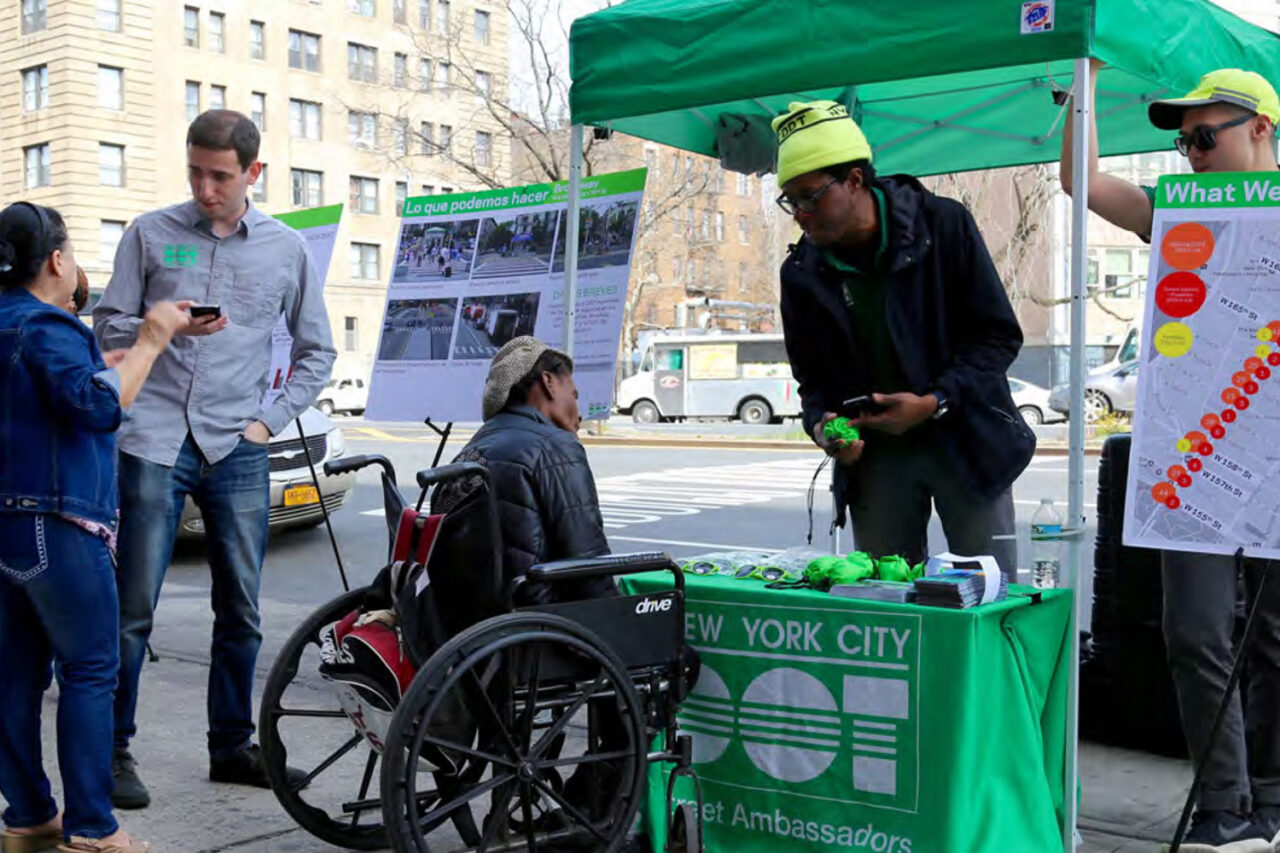 A man in a wheelchair speaking with a person an at information booth with a green awning and accessories such as sunglasses on dislay