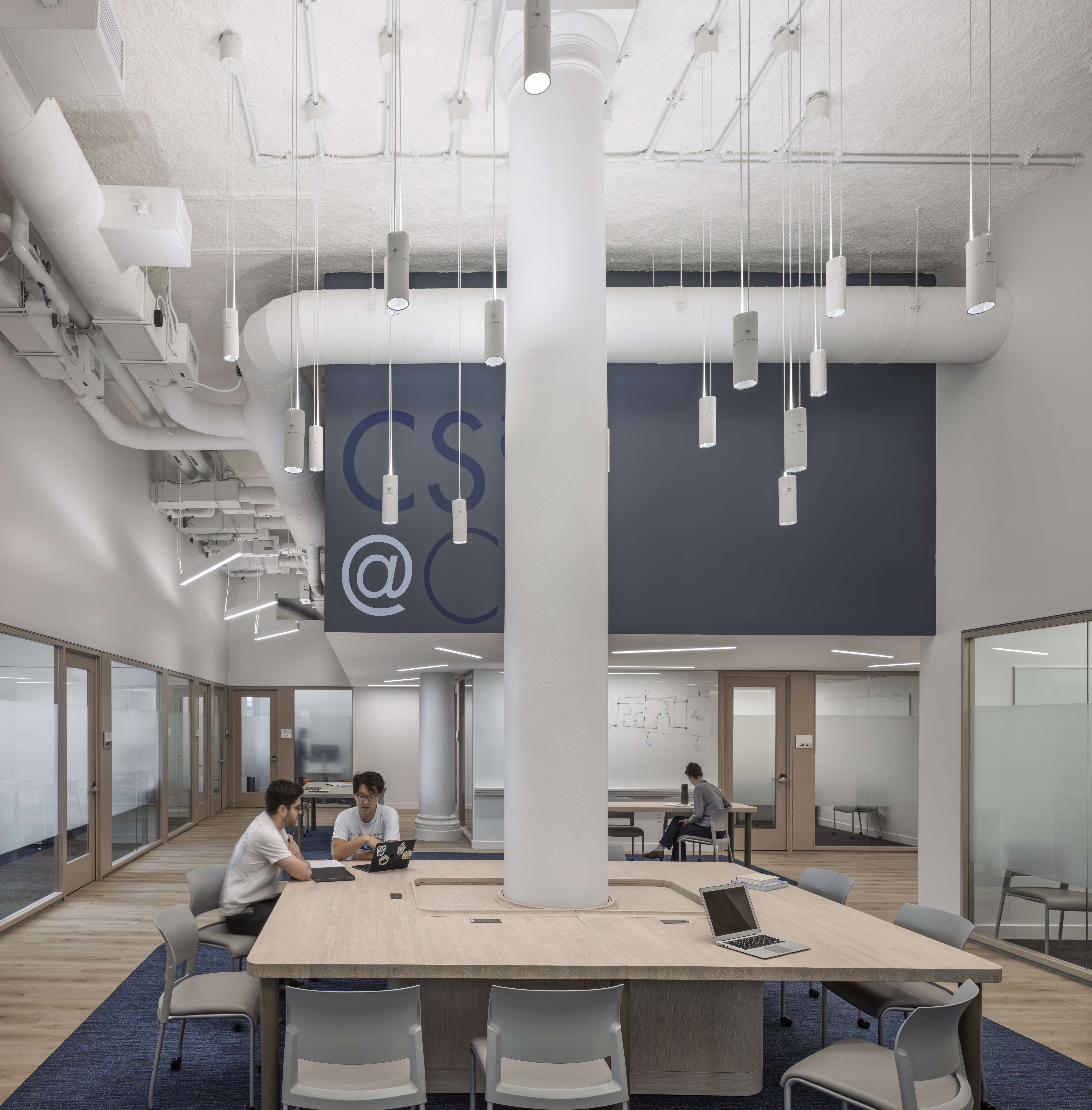Interior of the Columbia School of Engineering Computer Science Student Learning Center, showing students seated around a table and chairs with a tall white structural column in the center leading up through a double height space and exposed ceiling all in white