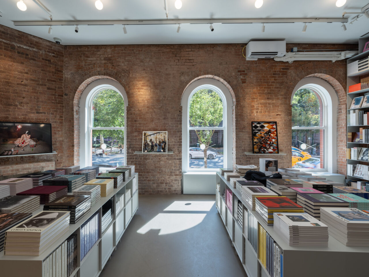Interior of the new Aperture space on Columbus Avenue and 78th Street, with stacks of books in the foreground and windows looking at a tree-lined streetscape