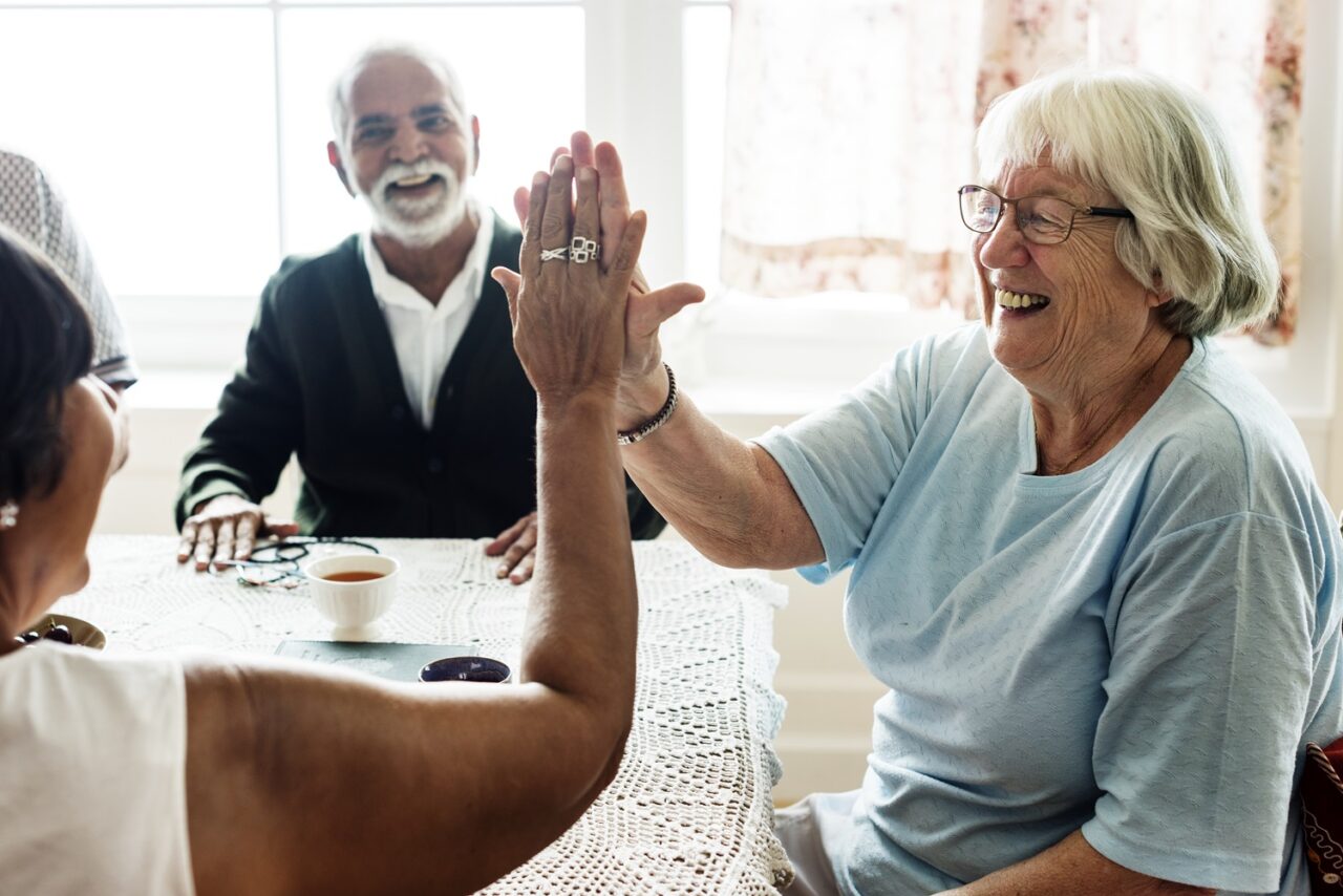 Two elderly women seated and doing a high five with an elderly man in the background