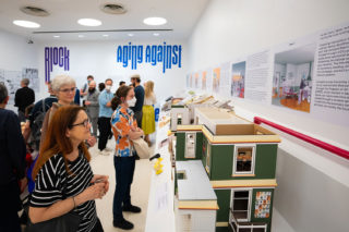 A crowd looks at architecture models and drawings in a gallery space.